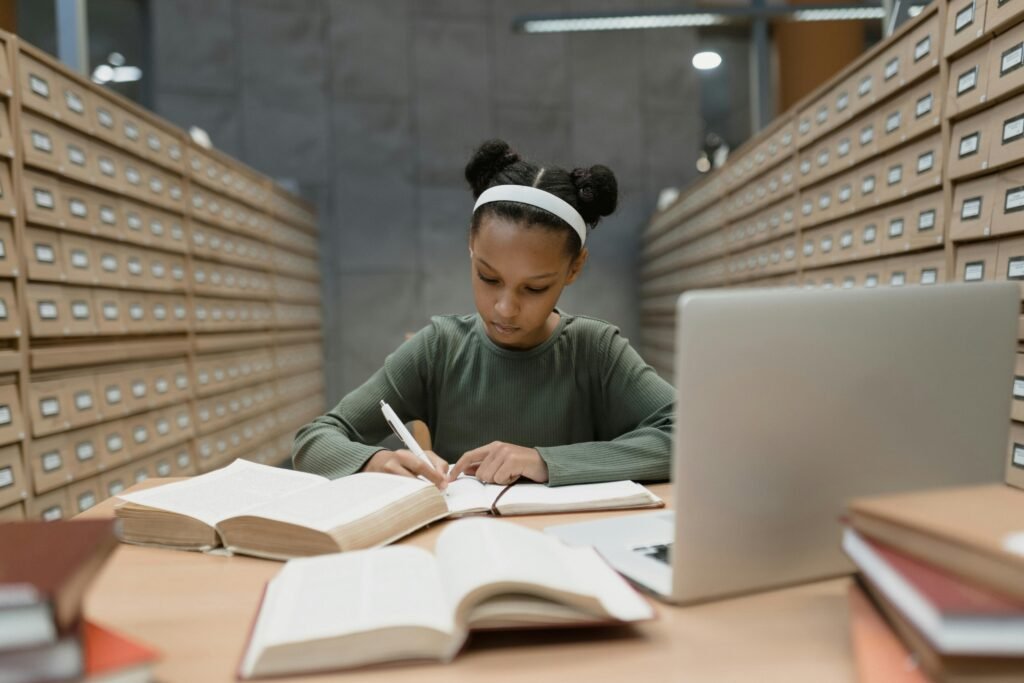 Jovem negra de cabelos presos em dois coques, usando fones de ouvido brancos e blazer cinza, concentrada estudando com livros abertos e um notebook em uma biblioteca. Ela está escrevendo em um caderno.